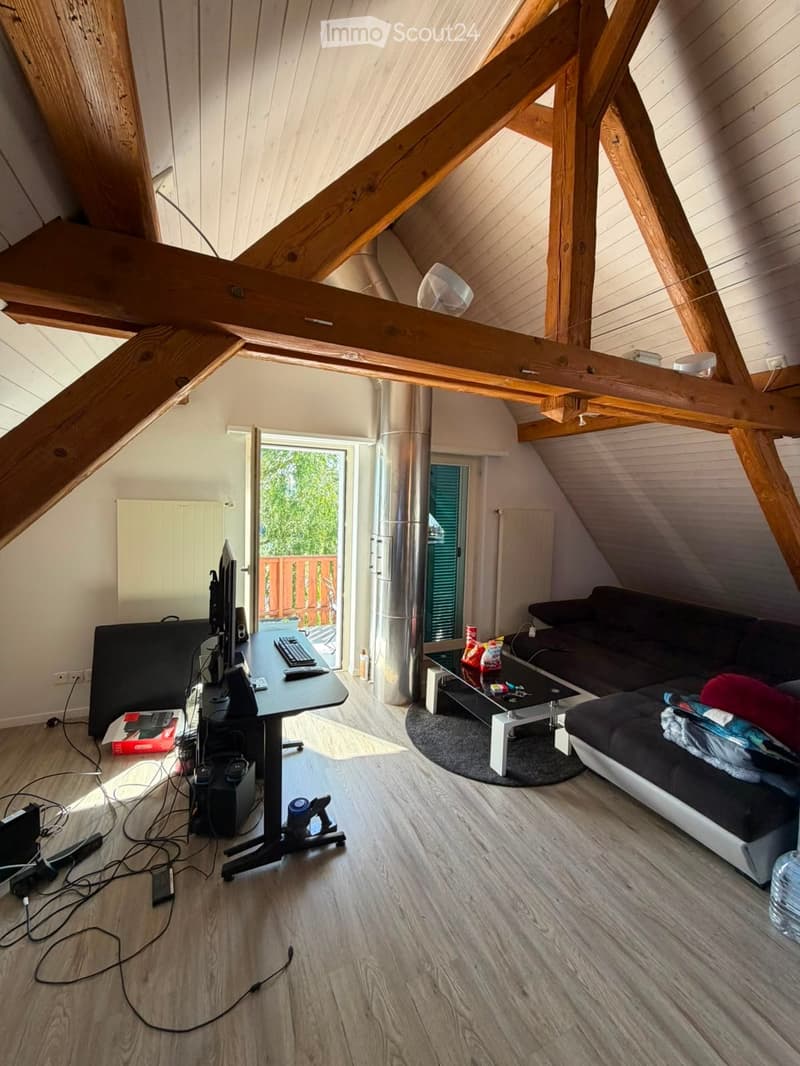 Loft-style living room with wooden beams, exposed ceiling, light wooden flooring, sofa, coffee table, desk with computer, and a view of the garden through a large window.