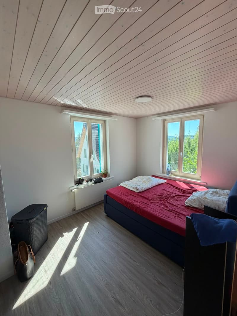 Bright bedroom with wooden floor, slanted wooden ceiling, large windows, bed with red and blue bedding, radiator, wicker basket, laptop, handbag.