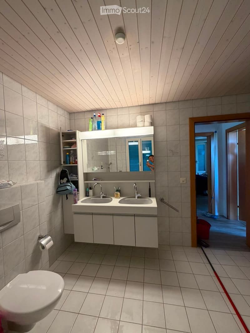 Bathroom with tiled walls and floor, double sink vanity with mirror, toilet, and various toiletries on the counter and shelves.