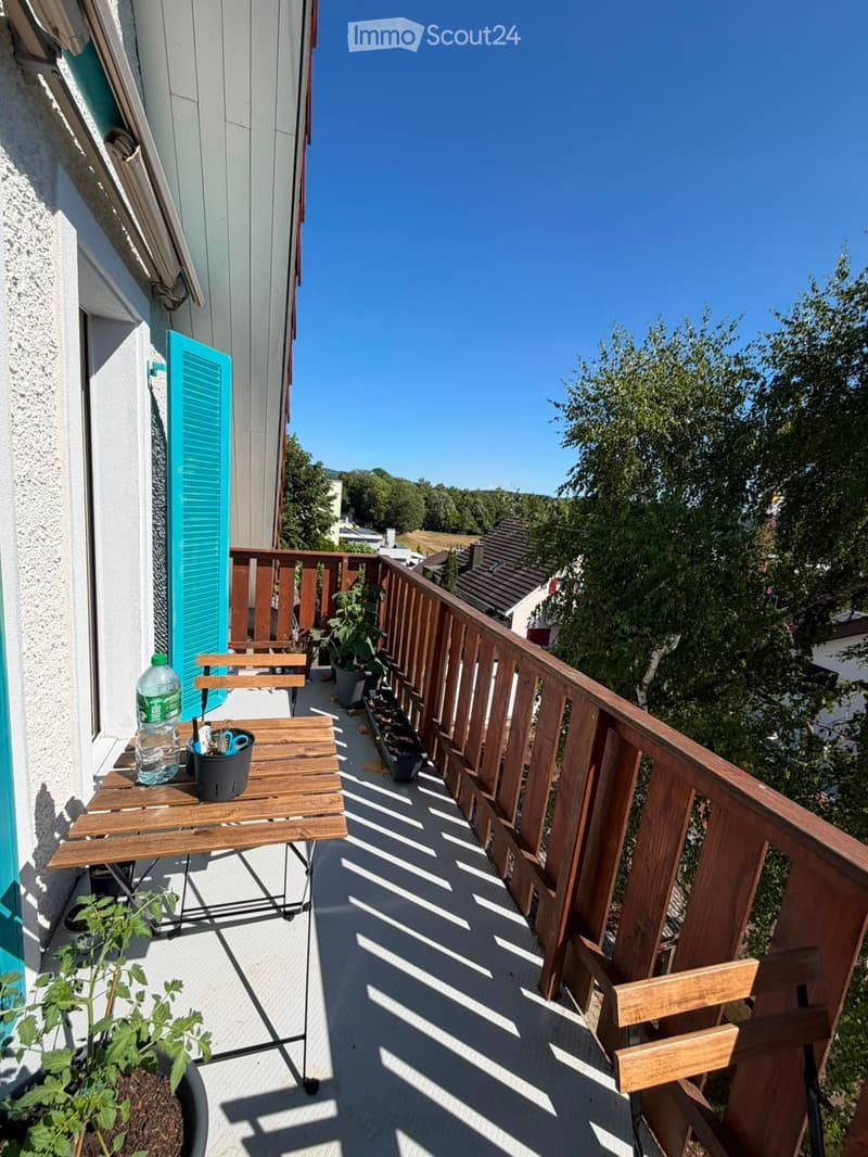 Balcony with wooden railing, small table, chairs, potted plants, and a bottle on the table.
