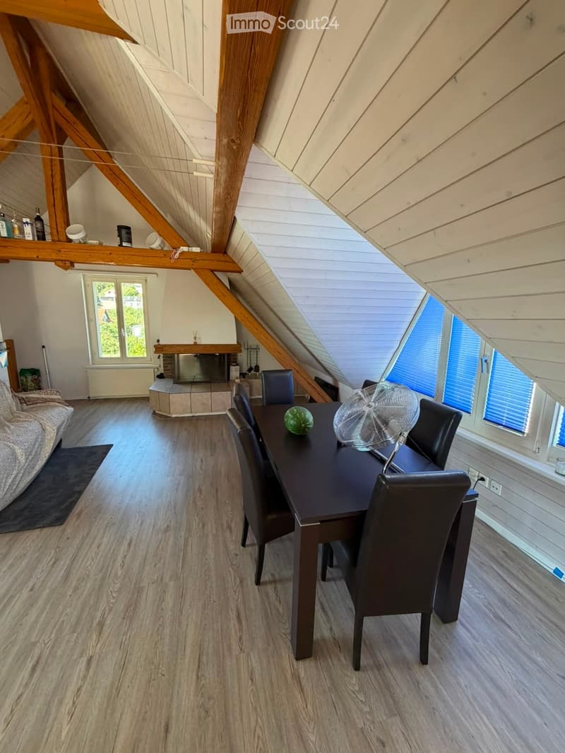 Loft-style room with wooden beams, fireplace, sofa, dining table with chairs, and a fan. Large windows provide natural light.
