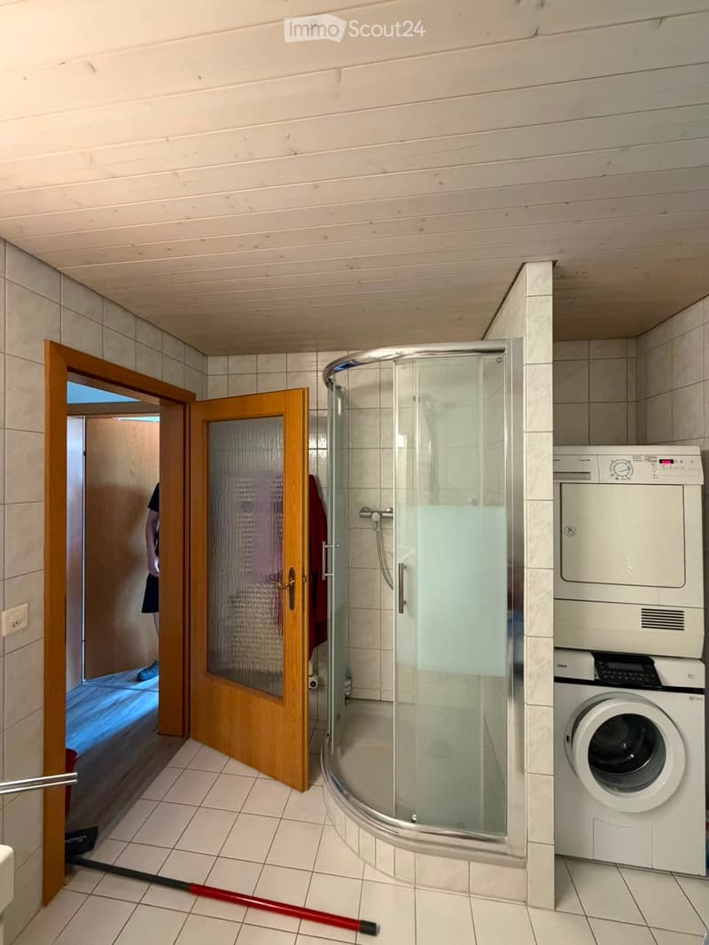 Bathroom with a glass-enclosed shower, a washing machine, and a wooden door leading to another room.