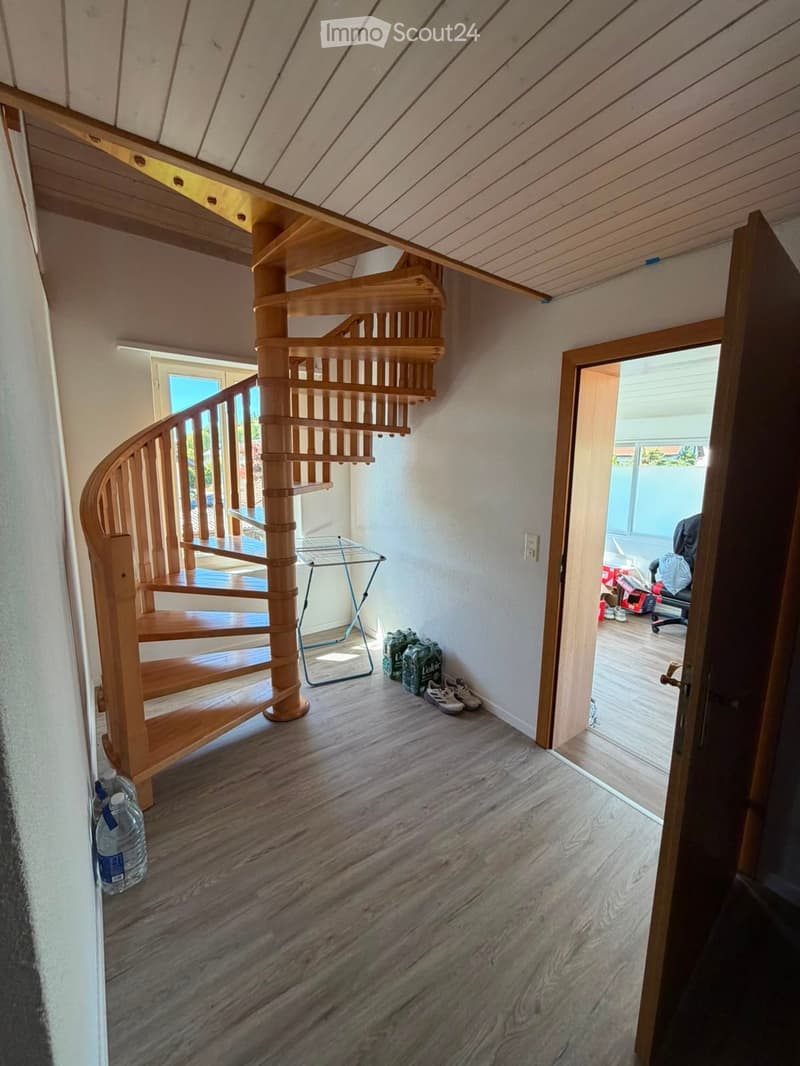 Spacious hallway with wooden flooring, a wooden spiral staircase leading upwards, and a small metal table with items on it. A doorway leads to another room with visible furniture and items.