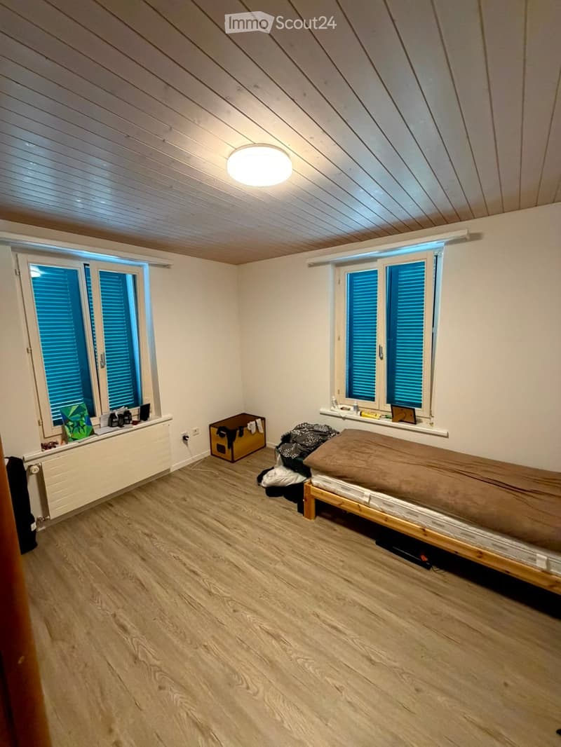 Cozy attic bedroom with wooden floors, slanted wooden ceiling, and large windows with blinds. Features a single bed, radiator, and storage box.