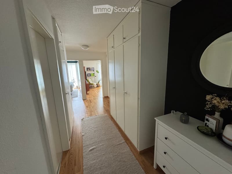 A well-lit hallway with wooden flooring, white walls, and a grey rug. A small table with decorative items is visible on the right side.
