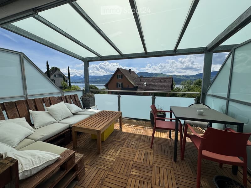 A modern, glass-enclosed balcony with wooden decking, a wooden sofa with white cushions, a wooden coffee table, and a small dining area with a table and chairs.