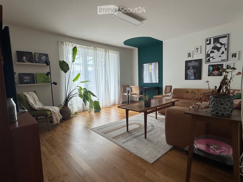 Modern living room with wooden flooring, a green accent wall, large windows with white curtains, a beige sofa, a wooden coffee table, a green armchair, and decorative shelves with artwork.