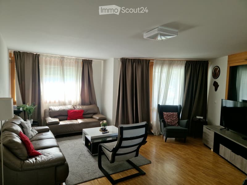 Modern living room with light-colored leather sofas, a coffee table, a TV, and a floor lamp. The room features wooden flooring, large windows with curtains, and decorative pillows on the seating.