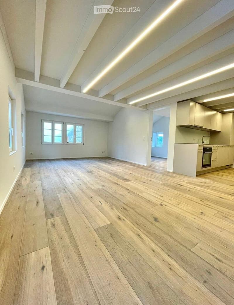 Empty room with wooden flooring, white walls, and exposed wooden beams on the ceiling. The room has large windows and an open doorway leading to a small kitchen area.