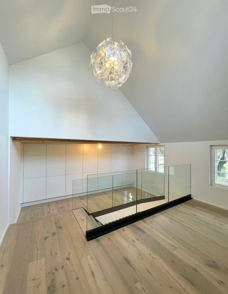 An empty room with wooden flooring, white walls, and a modern glass railing leading to a raised platform. The room features built-in white cabinetry and a large, modern chandelier hanging from the sloped ceiling.
