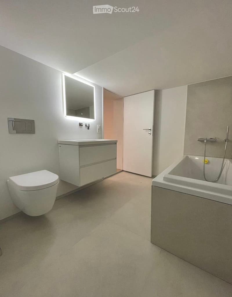 Modern bathroom with a toilet, sink, bathtub, and illuminated mirror. The space features light-colored tiles and sleek fixtures.