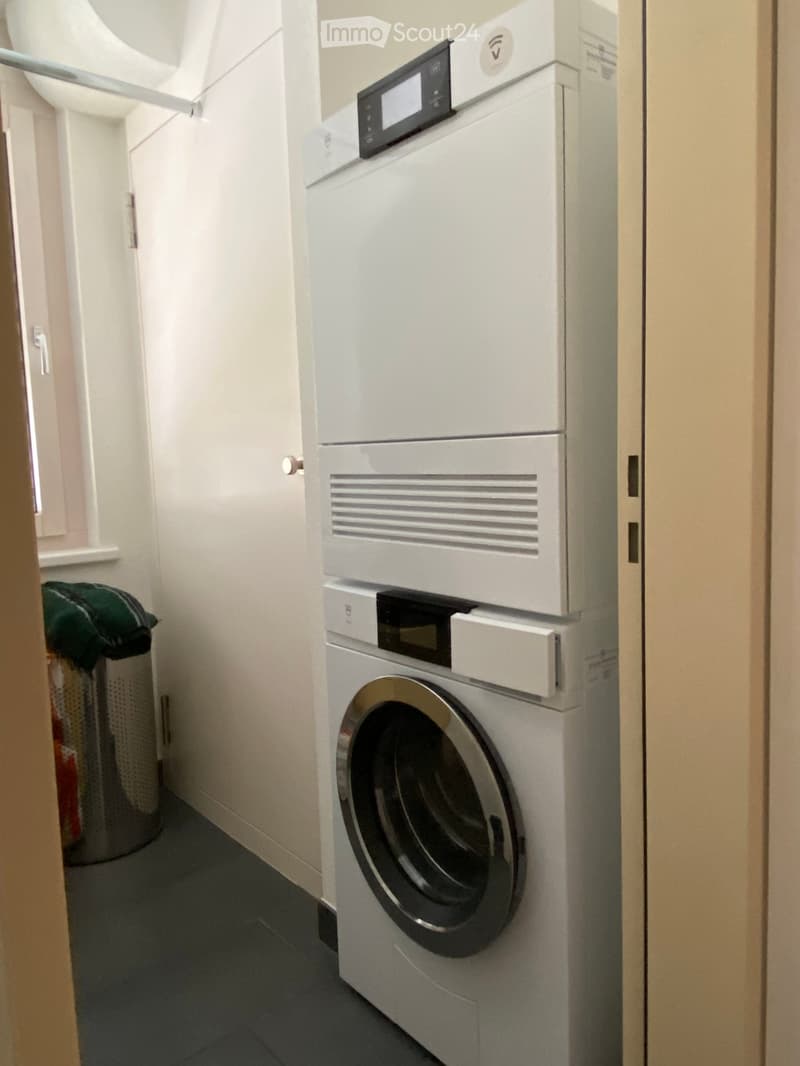 A compact bathroom with a stacked washing machine and dryer unit, a trash bin, and light-colored walls.