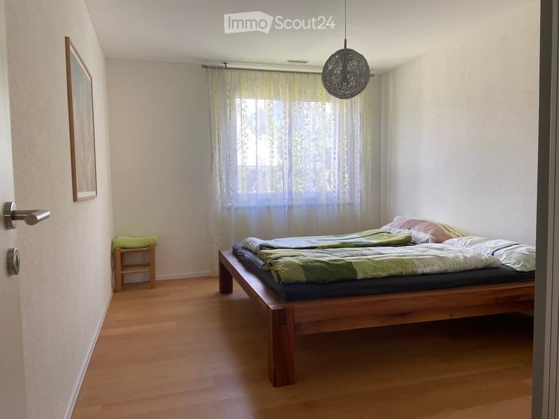 Bedroom with wooden floor, large window with sheer curtains, bed with green and white bedding, small wooden stool, and a mirror on the wall.