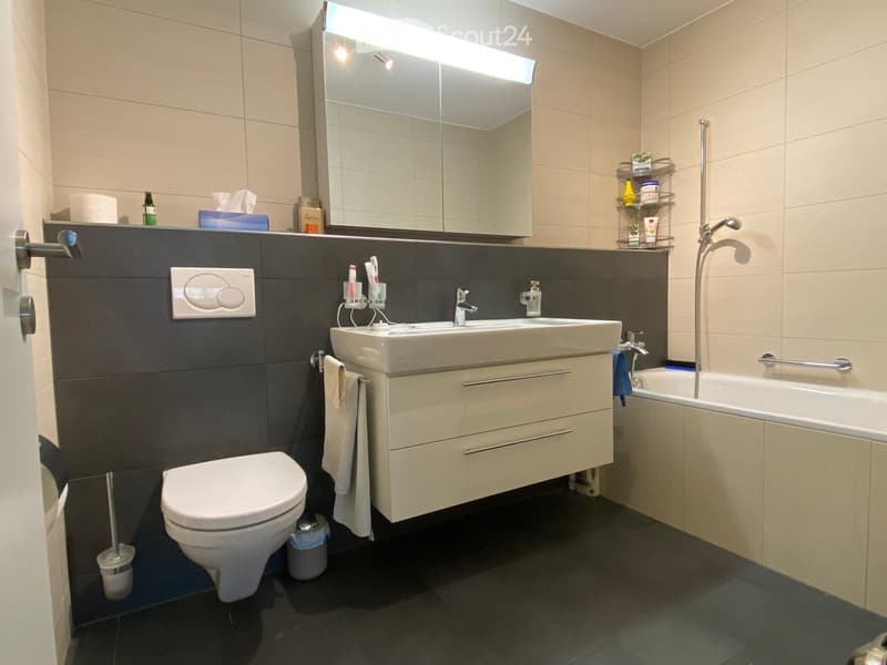 Modern bathroom with a white toilet, double sink, bathtub, large mirror, and various toiletries. Features dark tiles and a sleek design.