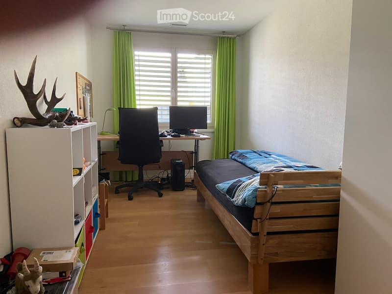 A small, well-lit bedroom with a wooden bed, desk, chair, and computer. The room features a window with green curtains, a shelf with decorative items, and a wooden floor.