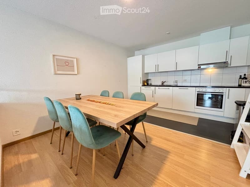 A modern, open-plan dining and kitchen area with a wooden dining table, teal chairs, white cabinets, and a built-in oven.