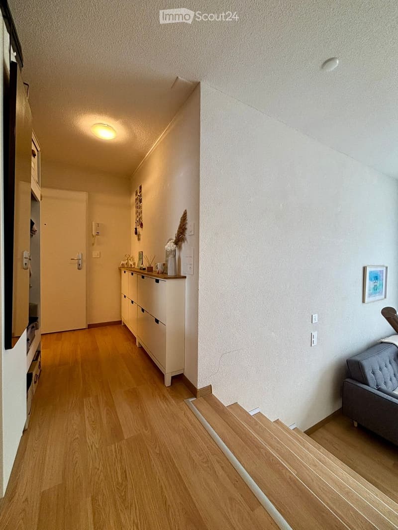 A narrow hallway with wooden flooring, white walls, and recessed lighting. Features a small cabinet with drawers, a vase with dried flowers, and a grey upholstered chair at the end.
