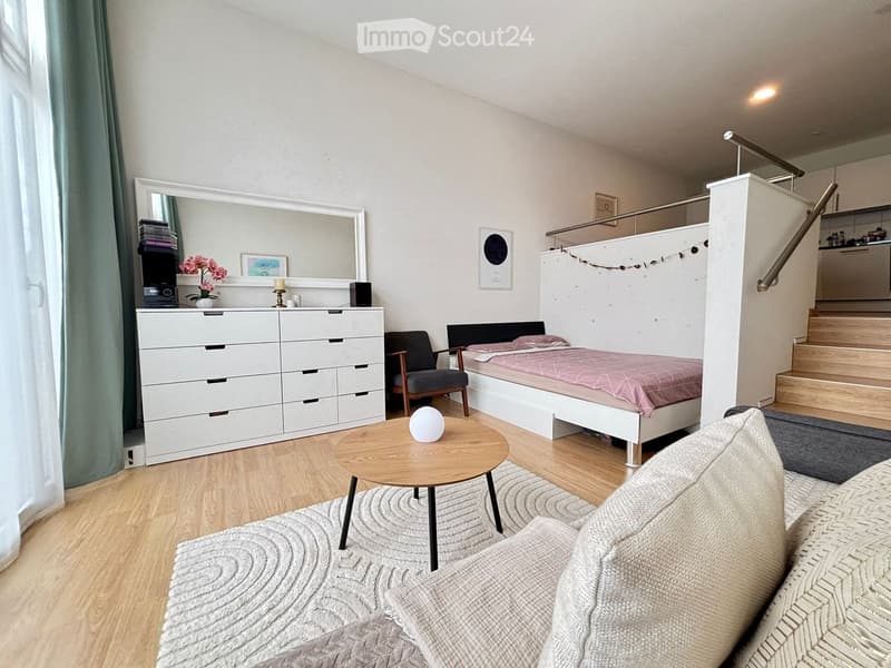 A modern, multi-functional room with a sofa, a small bed, a round wooden table, a chair, and a drawer unit. The space features a staircase leading to an upper level.