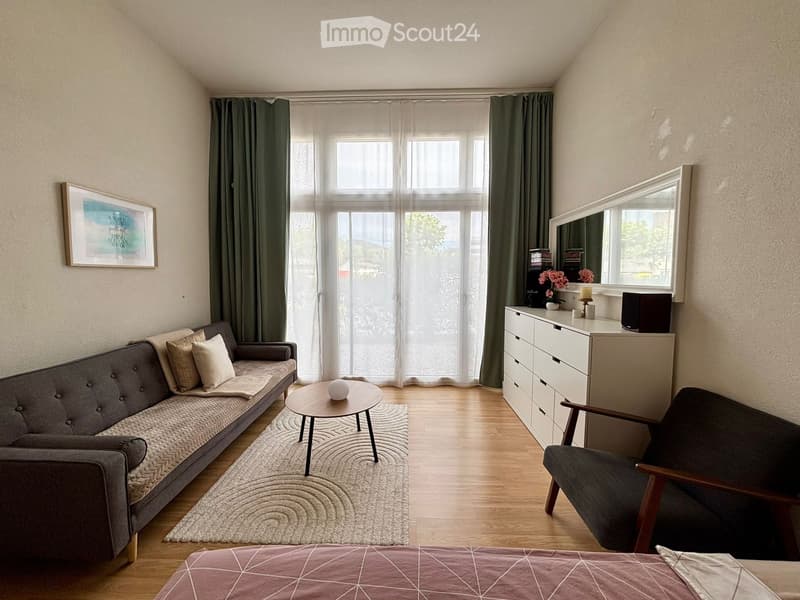 A modern living room with a grey sofa, wooden coffee table, armchair, white cabinet, and a large mirror. The room features large windows with green curtains and a light-colored rug.