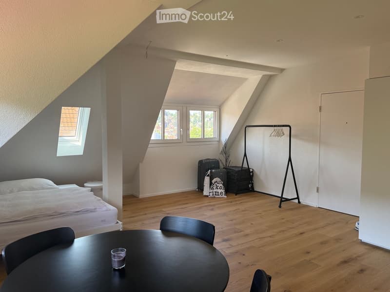 Bright attic bedroom with wooden flooring, slanted ceilings, and large windows. Features a bed, round table with chairs, and a coat rack.