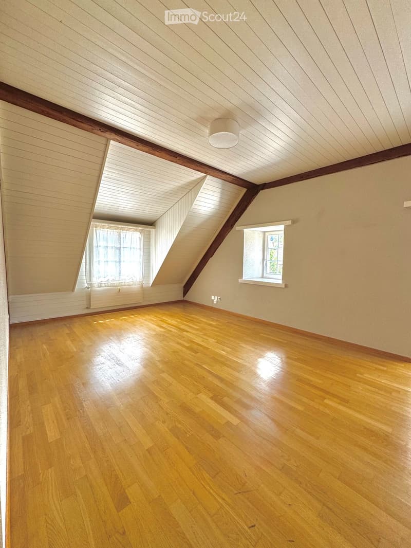 Empty attic room with wooden floors, slanted ceilings, and two windows, one with a curtain.