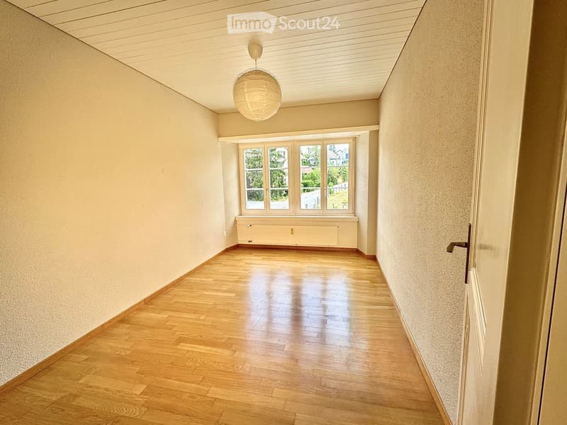 Empty hallway with wooden flooring, light-colored walls, and a large window at the end. A single hanging light fixture is visible.