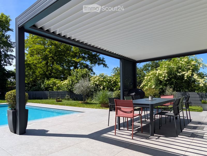 Covered outdoor patio with a dining table and chairs, surrounded by a well-maintained garden, a swimming pool, and lush greenery.