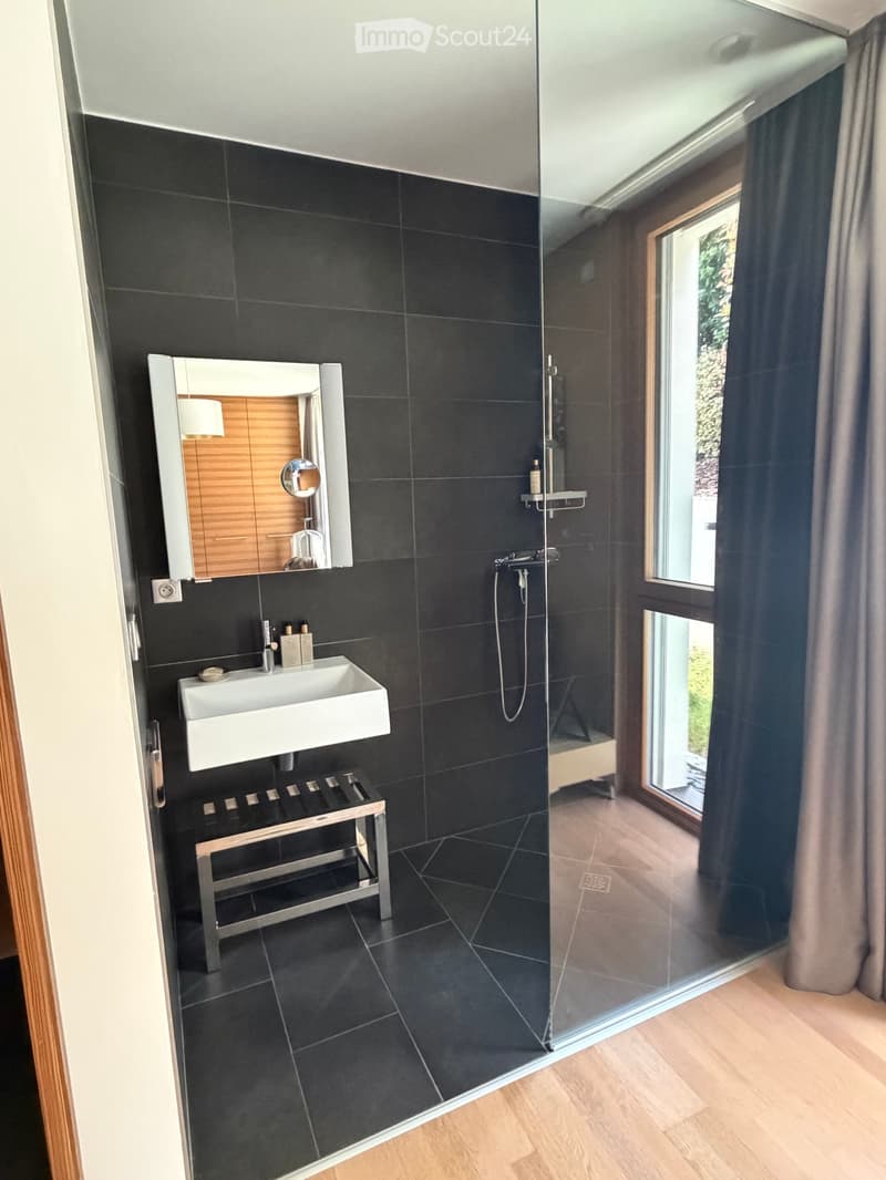 Modern bathroom with black tiles, white sink, mirror, glass shower enclosure, and a small stool.
