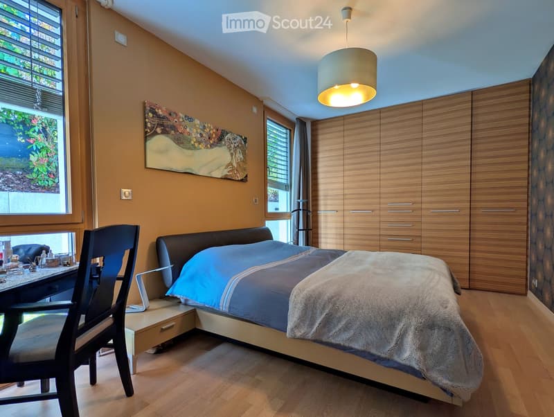 Modern bedroom with a large bed, wooden wardrobe, a chair, a small table, and a hanging lamp. The room features wooden flooring and large windows with blinds.