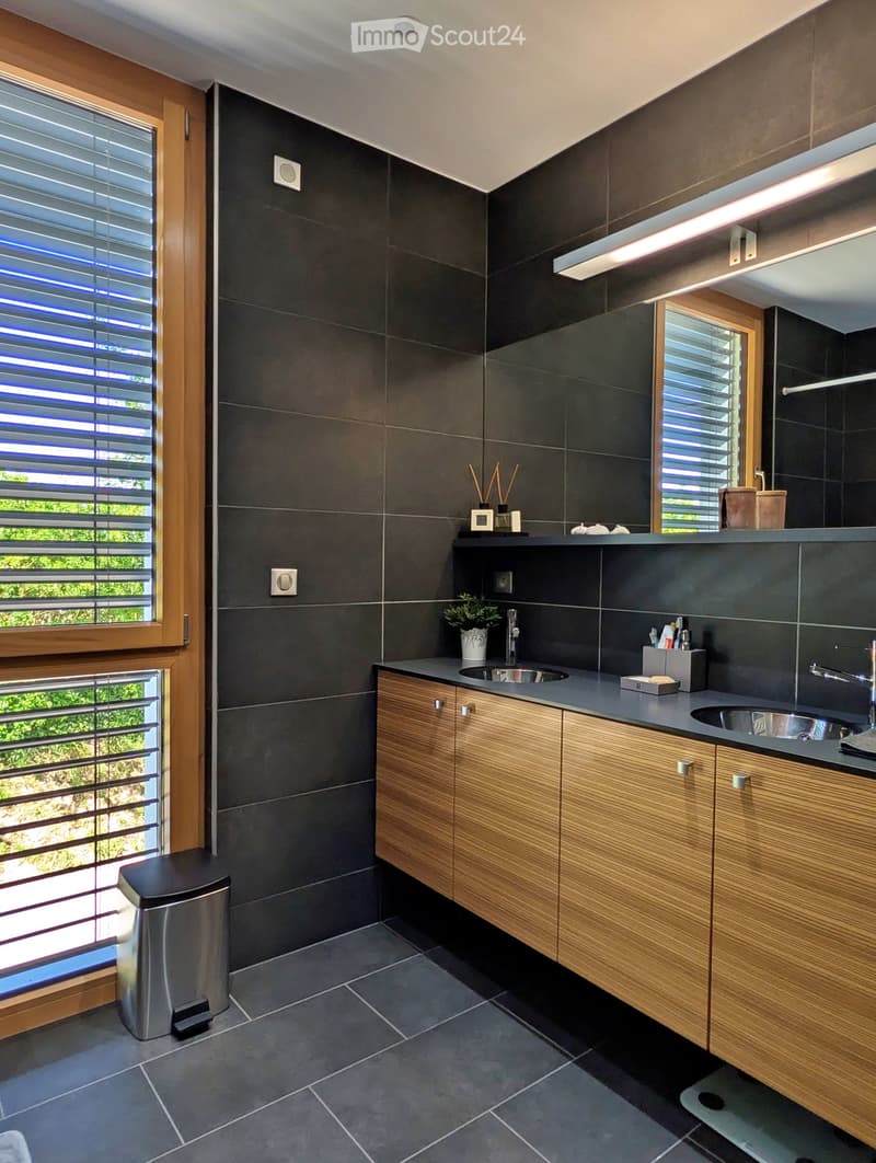 Modern bathroom with dark grey tiles, wooden cabinets, double sink, and large windows with blinds.