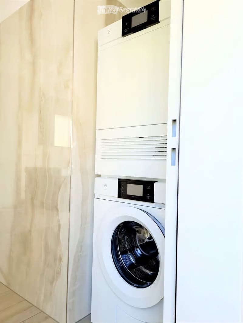 Modern bathroom with a white washing machine and dryer stacked vertically, featuring a sleek digital control panel and a light-colored tiled wall.