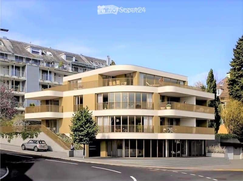 Modern multi-story residential building with large windows, balconies, and contemporary design. The building features a combination of beige and brown tones, with a sleek, curved facade and multiple levels.