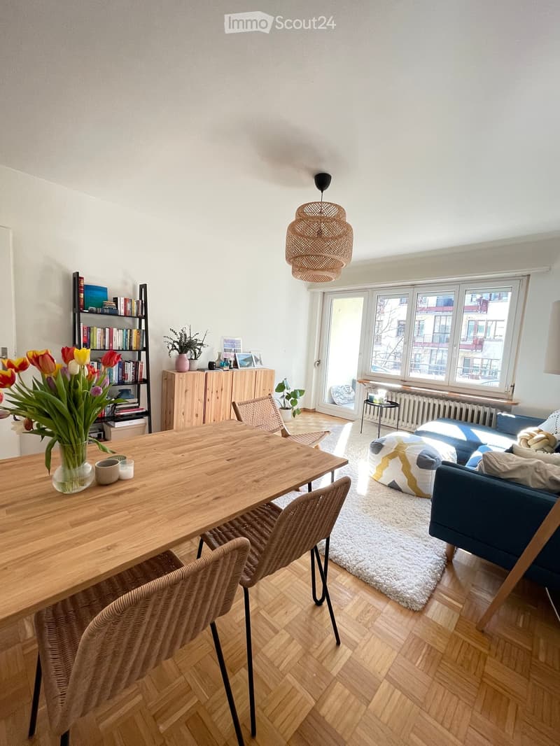 Bright living room with wooden flooring, large windows, a blue sofa, a wooden dining table with chairs, a bookshelf, and decorative items.
