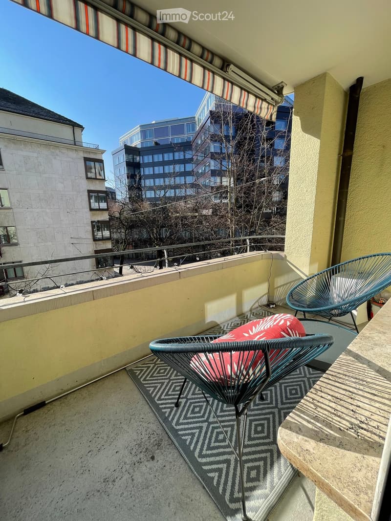 Balcony with modern chairs, a small table, and a patterned rug, overlooking a cityscape with modern buildings.