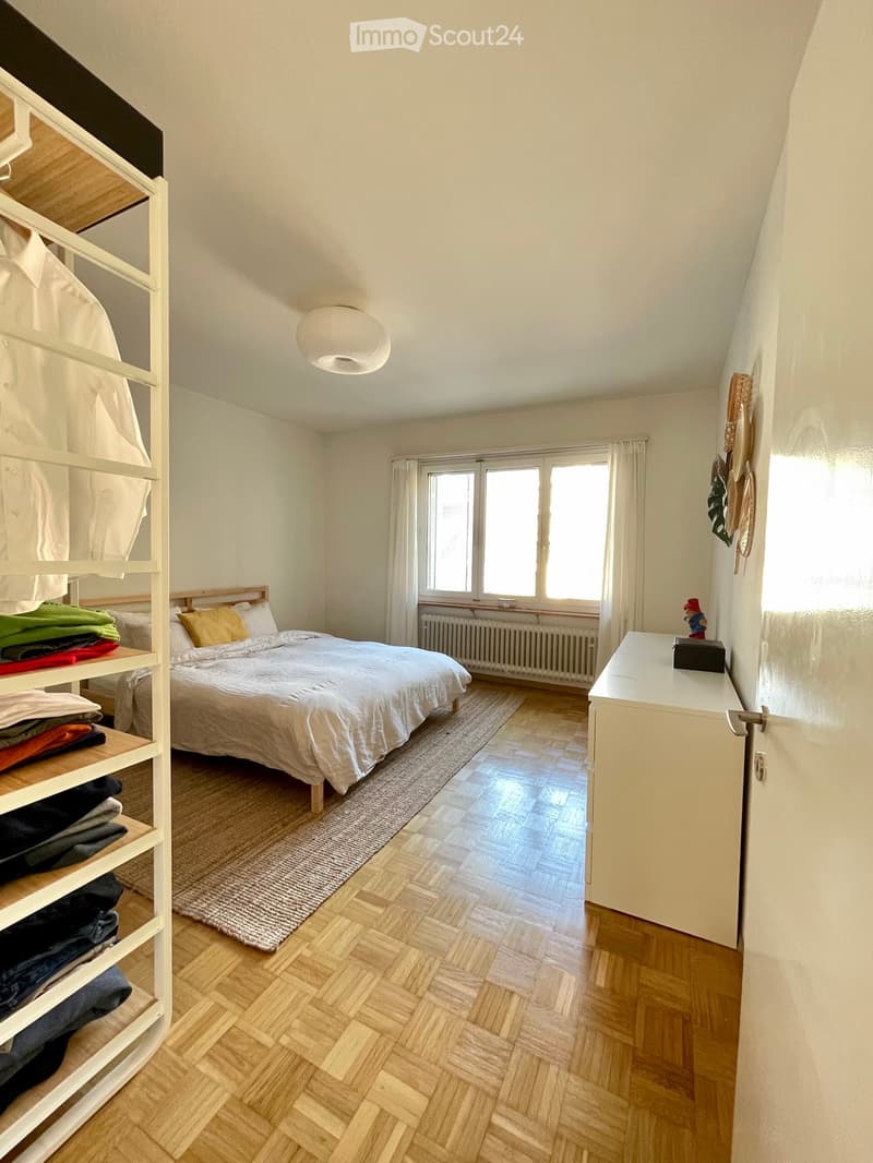 Bright bedroom with wooden herringbone flooring, a double bed with white bedding, a nightstand, a shelf with folded clothes, and a radiator under the windows.