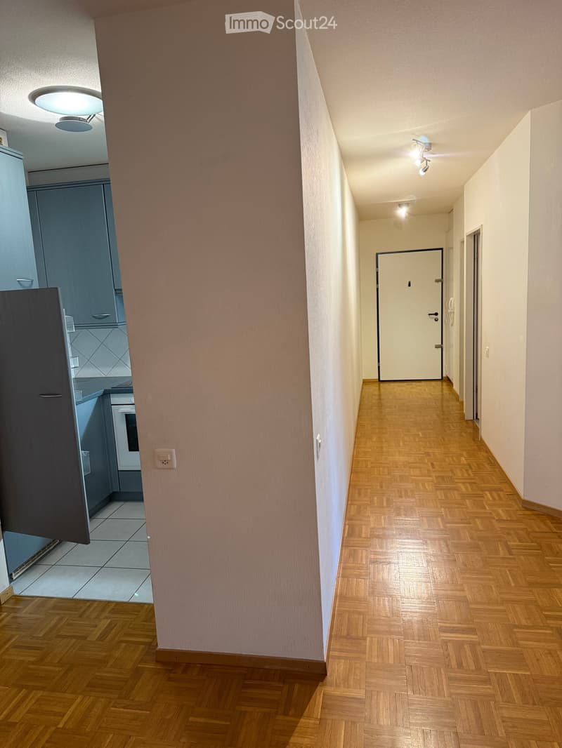 A narrow hallway with wooden flooring, light-colored walls, and recessed ceiling lights. A door is visible at the end of the hallway, and part of a kitchen area is visible on the left side.