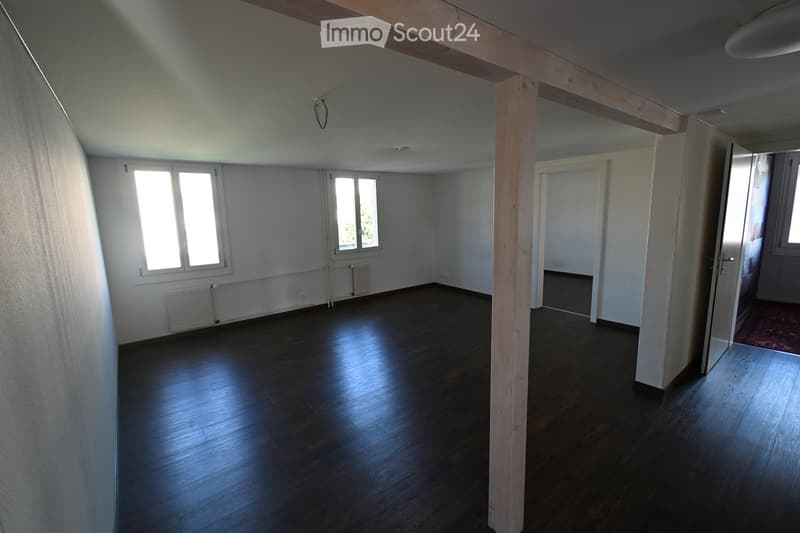 Empty room with wooden flooring, white walls, and large windows allowing natural light. There are radiators along the walls and a wooden support beam in the center.