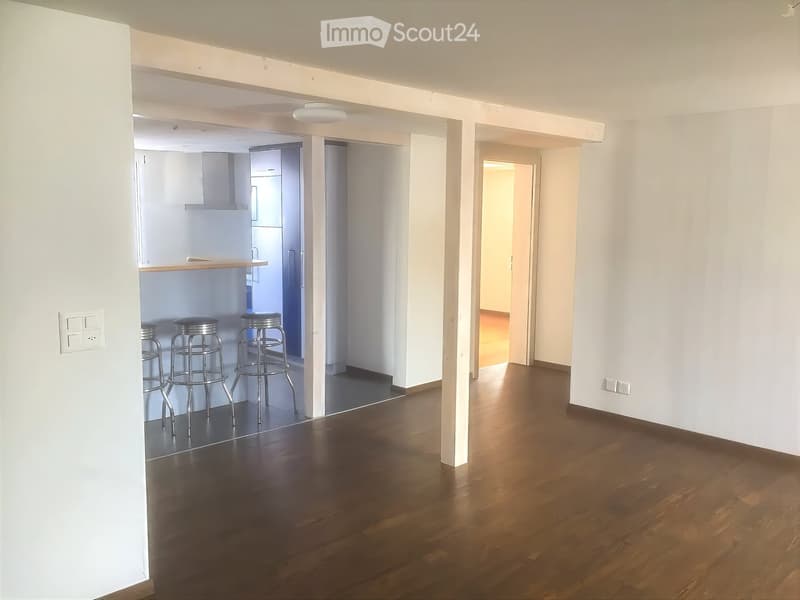 Open-plan living and dining area with wooden flooring, white walls, and four stools near a kitchen counter.