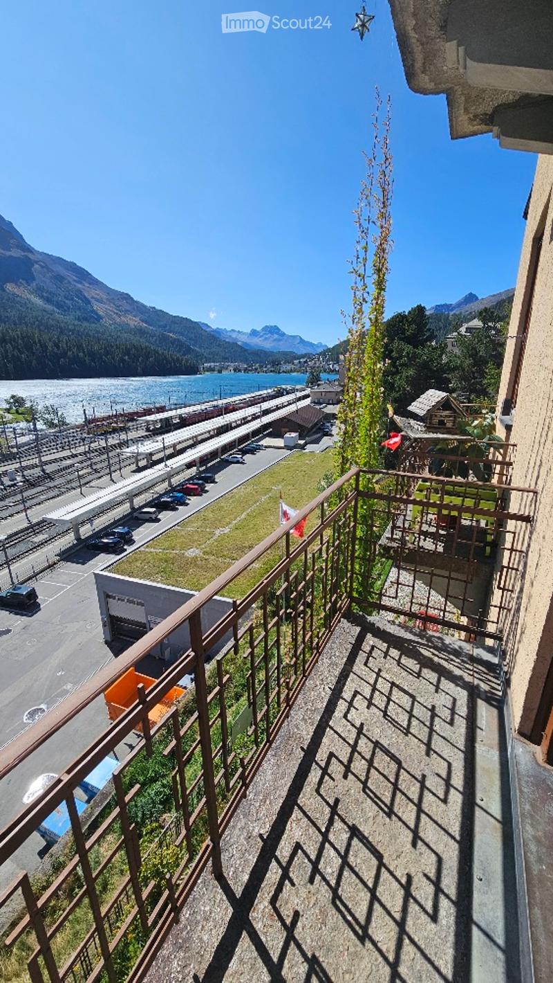 Balcony with metal railing overlooking a scenic view of mountains, a river, and a town below. The area includes a small garden patch and a few parked cars in the distance.