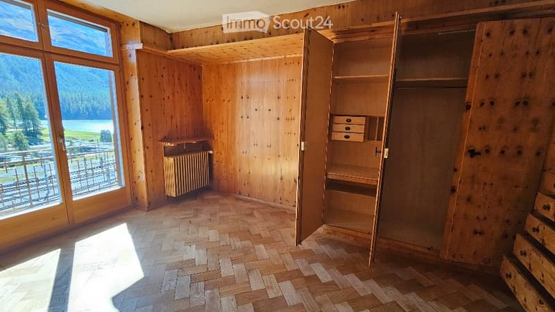 Empty room with wooden walls, herringbone wooden flooring, large windows with scenic mountain view, built-in closet with shelves and drawers, radiator, and balcony access.