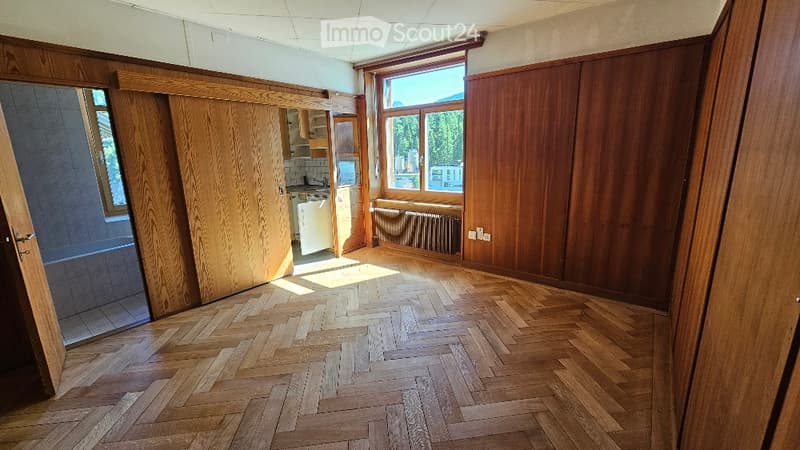 Empty room with herringbone wooden flooring, large windows, and wooden paneling on walls. Adjacent kitchen area visible through an open doorway.