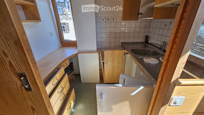 The kitchen features light wooden cabinets, a sink with a faucet, and a countertop with a plate. The space is well-lit with natural light coming through a window, and the floor is a neutral gray color.
