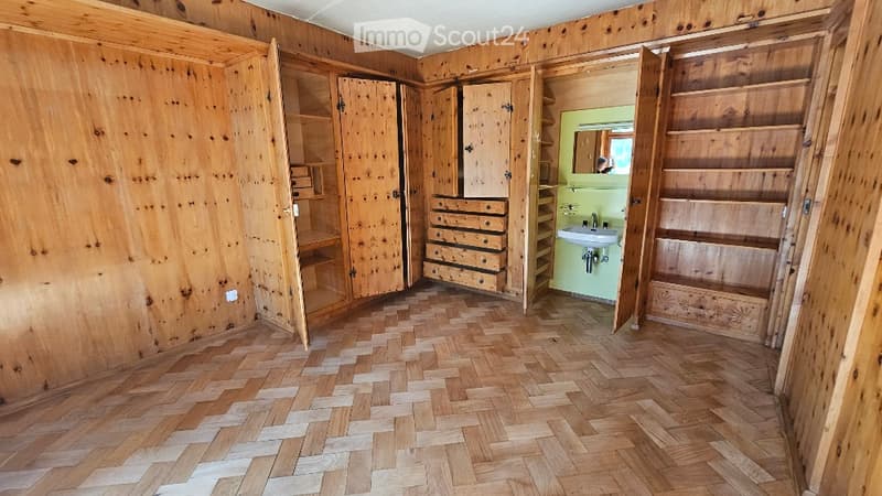 Empty room with wooden walls, herringbone wooden flooring, and built-in wooden cabinets and drawers. A small adjacent bathroom with a sink and mirror is visible.