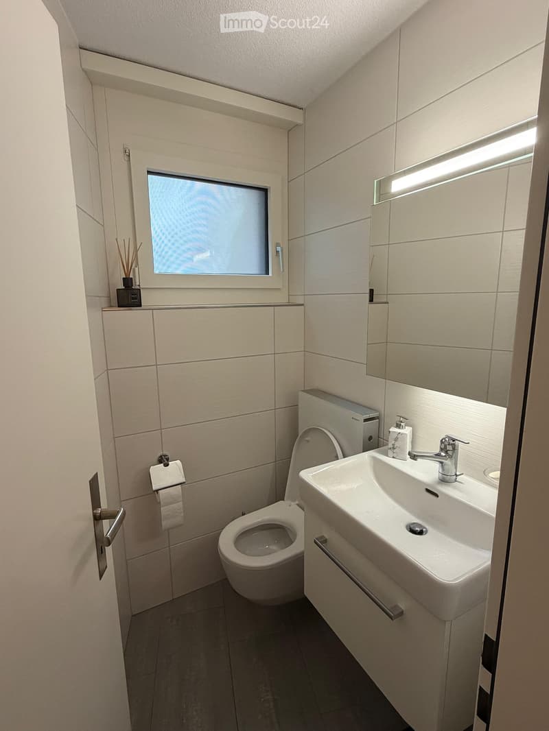 Modern bathroom with white tiles, a toilet, a sink with a modern faucet, a mirror, and a small window with frosted glass. Includes a soap dispenser and toilet paper holder.