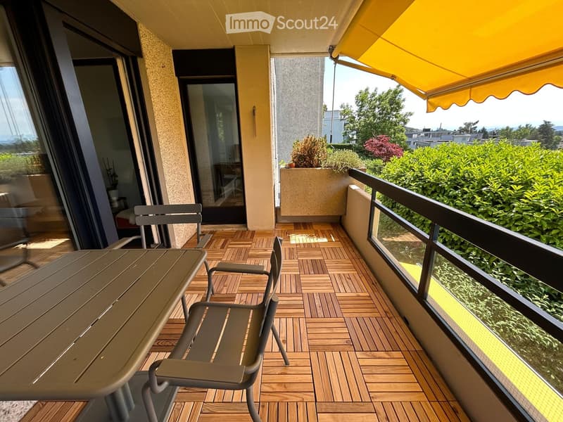 Balcony with wooden decking, table, chairs, and a yellow awning, overlooking greenery and a cityscape.