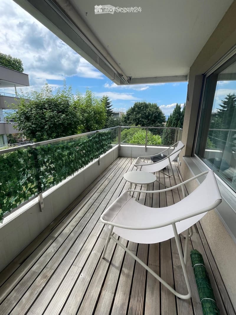 Balcony with wooden decking, two white chairs, a small round table, and a glass railing with greenery visible beyond.