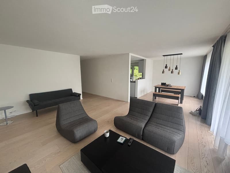 Modern living room with light wooden flooring, grey sectional sofa, black coffee table, and a dining area with a wooden table and benches.