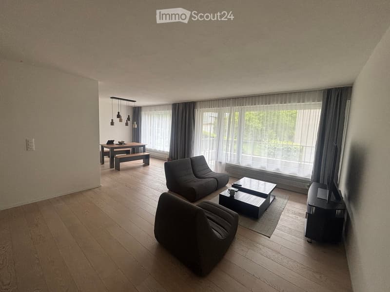 Modern living room with wooden flooring, large windows, grey sofa, coffee table, dining table, and bench.