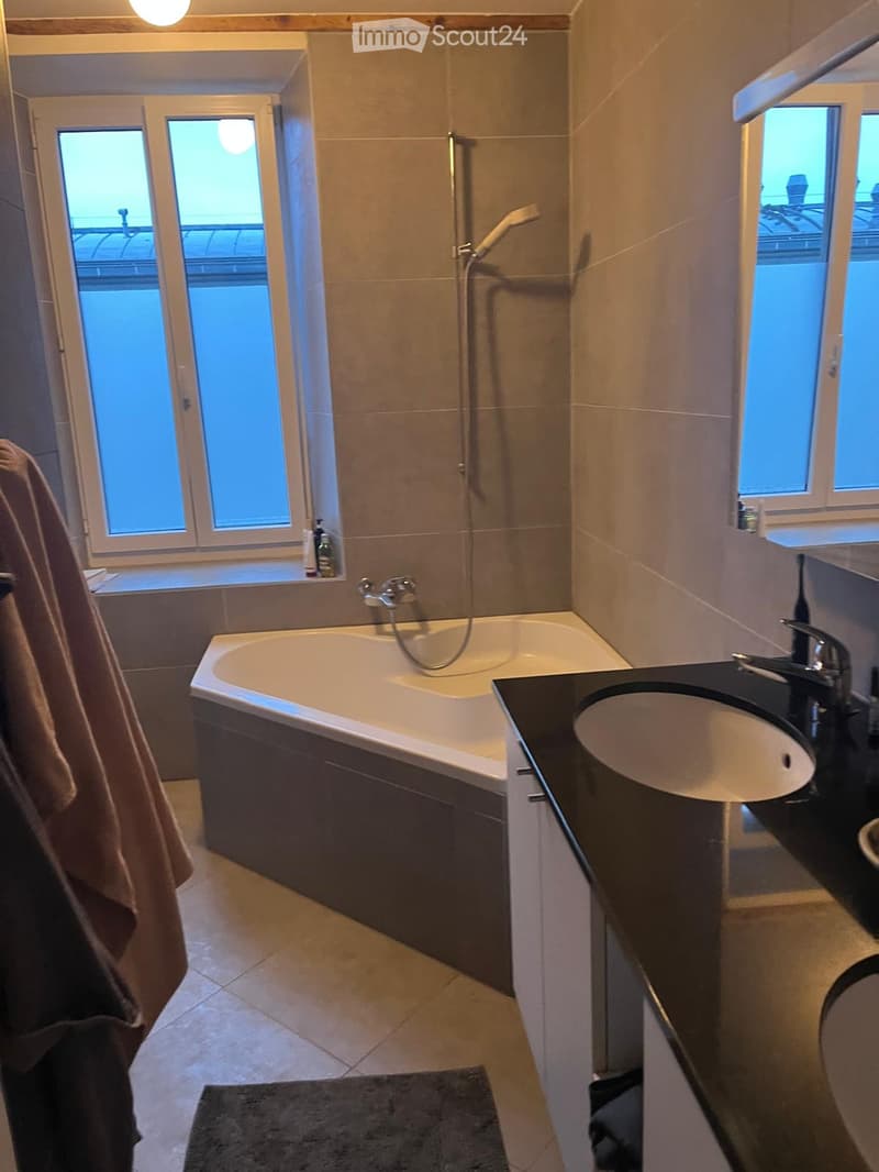 Modern bathroom with a bathtub, sink, showerhead, and towel visible. The room features large windows and a sleek design.