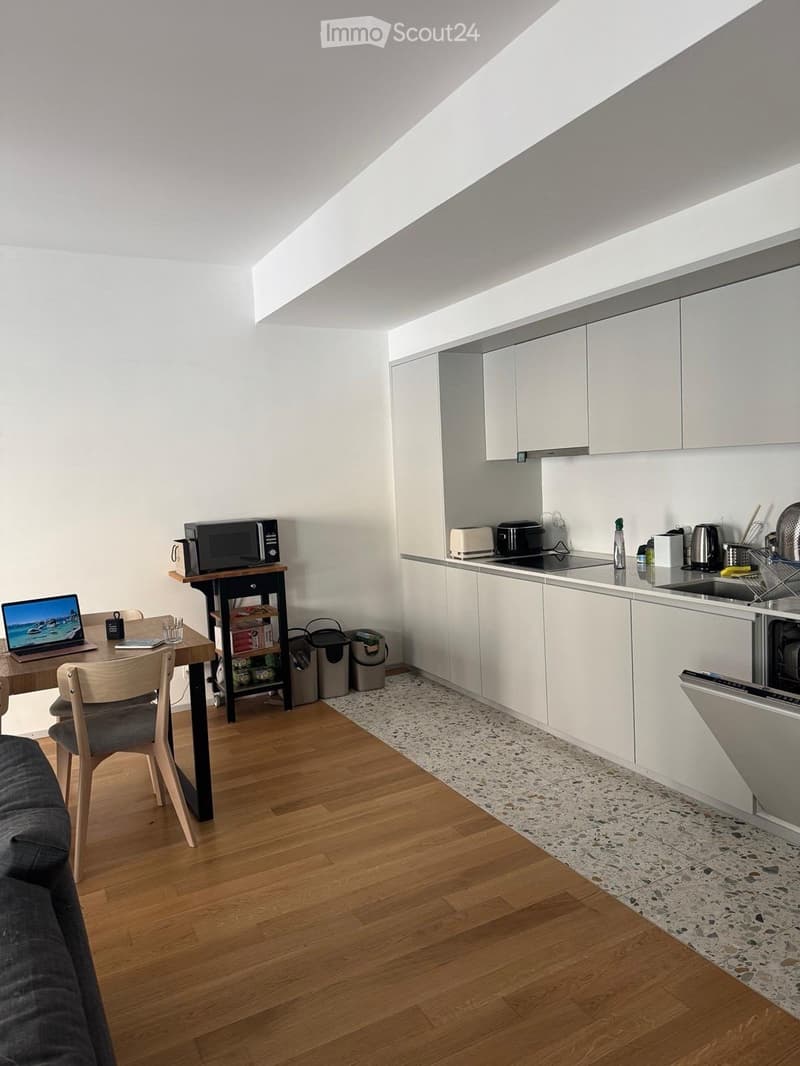 A modern, open-plan kitchen and dining area with white cabinets, a wooden dining table, and various appliances including a microwave, toaster, and dishwasher.